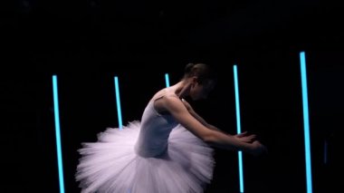 Kamera, beyaz etekli, klasik balerin unsurlarıyla şehvetli balerin etrafında dönüyor. Beyaz kuğu yavaş çekimde parlak neon ışıklarla siyah stüdyonun arka planına doğru hareket ediyor. Kapat.