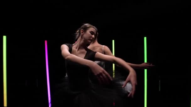 Φωτογραφική κυκλική κίνηση γύρω από νεαρές μπαλαρίνες που χορεύουν σε μαύρο φόντο στούντιο με φωτεινά φώτα νέον. Κομψές κινήσεις ενός ασπρόμαυρου κύκνου σε αργή κίνηση. Τροχιακή βολή από κοντά. — Αρχείο Βίντεο