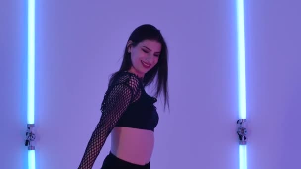 Tânăra femeie drăguță dansând benzi în studio întunecat pe fundalul luminilor luminoase de neon. Dansatoare de sex feminin coquettishly se uită la camera foto, demonstrează corpul ei sexy și plasticitatea. De aproape. Slow motion . — Videoclip de stoc