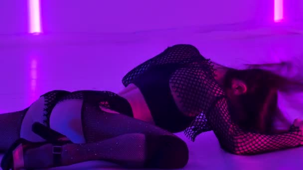 Sportieve jonge vrouw danst erotisch een strip terwijl ze op de vloer ligt in een donkere studio met fel neonlicht. De brunette demonstreert bindgaren en stretching. Sluit maar af. Langzame beweging. — Stockvideo
