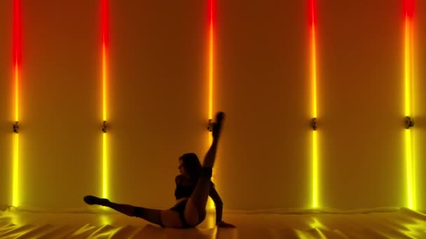 Sexy stripper erotically dances strip with elements of aerobics in dark studio against background of yellow red neon lights. Hot brunette plastic moves demonstrating her beautiful body. Slow motion. — Stock Video