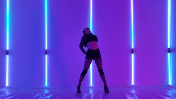 Horká brunetka eroticky tančí striptýz v tmavém studiu na pozadí jasných neonových světel. Striptérka se hravě dívá do kamery, demonstruje svou sexualitu a flexibilitu. Zpomalený pohyb. — Stock video
