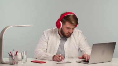 Öğrenci internetten büyük kırmızı kulaklıklarını çıkarıp gevşemesini izlemekten yoruldu. Gündelik giyinen genç adam ofis masasında otururken uzaktan öğrenme yapıyor. Kapatın. Yavaş çekim.