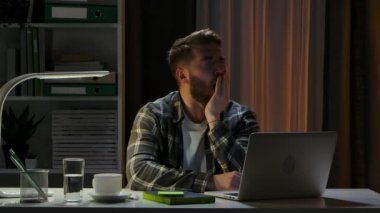 Yorgun bir adam evdeki ofis masasında dizüstü bilgisayarın önünde oturur ve esner. Evden uzak bir iş. İşte uzun bir günün sonu. Serbest çalışıyorum. Online eğitim. Kapatın. Yavaş çekim.