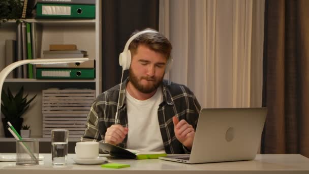Pozytywny brodaty człowiek pracujący zdalnie na laptopie, siedząc w biurze. Freelancer lubi przyjemną muzykę noszącą duże białe słuchawki. Nowa koncepcja pracy w domu Zbliżenie. Zwolniony ruch. — Wideo stockowe