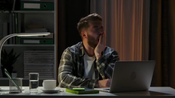 Unavený vyčerpaný muž sedí ve tmě u stolu v kanceláři před laptopem a zívá. Dálková práce z domova. Konec dlouhého dne v práci. Na volné noze. Online školení. Zavřít. Zpomalený pohyb. — Stock video