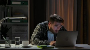 Dizüstü bilgisayarda klavyeye dikkatlice bakıp daktilo eden konsantre genç bir iş adamı. Freelancer karanlık bir ofiste otururken uzaktan çalışıyor. Kapatın. Yavaş çekim.