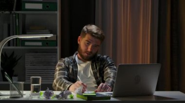Yorgun genç adam karanlık bir ofiste dizüstü bilgisayarın başında oturuyor. Freelancer yapraklar üzerine notlar alır ve onları sıkar, iç çeker, daha fazla üzgün fikir yoktur. Evden uzak bir iş. Kapatın. Yavaş çekim.