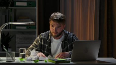 Yorgun genç adam karanlık bir ofiste dizüstü bilgisayarın başında oturuyor. Freelancer doğru fikri bulamadığı için üzgün bir şekilde yaprakları karıştırıyor. Evden uzak bir iş. Online eğitim. Kapatın. Yavaş çekim.