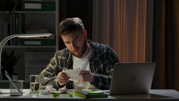 Unavený mladík sedí u laptopu v tmavé domácí kanceláři. Freelancer se dívá listím s poznámkami, je rád, že našel správný nápad. Dálková práce z domova. Zavřít. Zpomalený pohyb. — Stock video