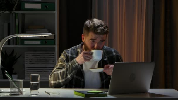 Kreatywny człowiek siedzący przy biurku w pracowni domowego biura, pracujący wieczorem nad laptopem. Zdalny pracownik pracuje i lubi gorącą kawę. Praca zdalna, trening online w domu. Zamknij drzwi. Zwolniony ruch. — Wideo stockowe