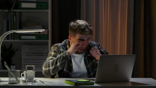 Unavený muž sedí a spí za stolem v domácím kancelářském studiu poblíž notebooku večer. Dálkový pracovník se probouzí, pije kávu a pokračuje v psaní na počítači. Vzdálená práce. Zavřít. Zpomalený pohyb — Stock video
