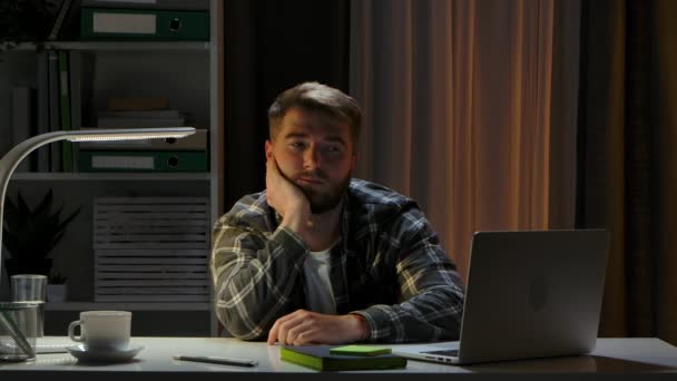 到了晚上，累了的男人坐在笔记本电脑旁的总公司工作室的办公桌前。那个遥远的工人在等着工作日的结束.远程工作，在家接受在线培训。靠近点慢动作. — 图库视频影像