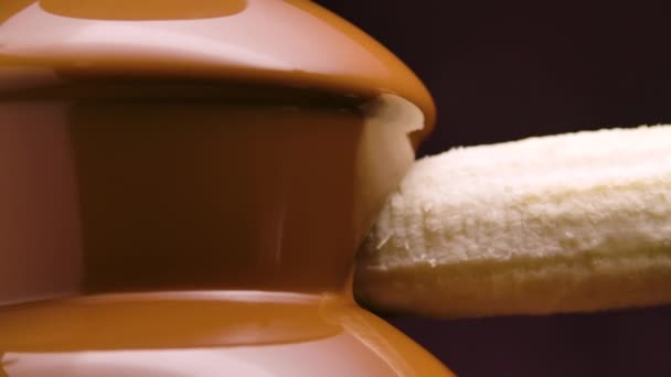 Melted milk chocolate flows in a chocolate fountain. Ripe banana is dipped and wrapped in hot liquid chocolate flowing down from the cascades. Fondue. Party and celebration. Close up. Slow motion. — Stock Video
