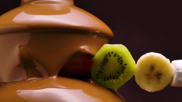 Ripe juicy fruits on skewer dipped in sweet melted chocolate fountain. Banana, kiwi, orange, marshmallows and strawberry wrapped in hot liquid chocolate. Fondue. Dessert. Close up. Slow motion. — Stock Video