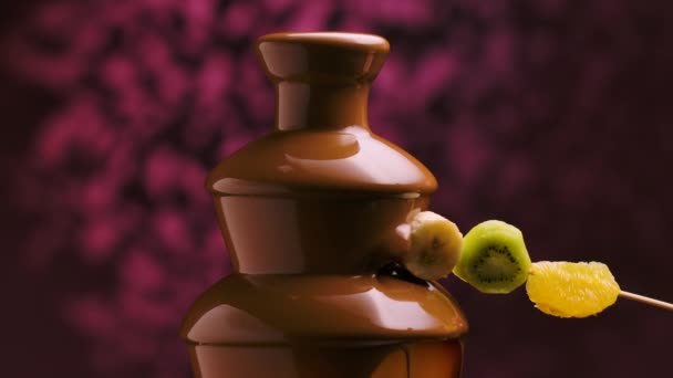 Mans mão mergulha um espeto com frutas em chocolate derretido doce na fonte de chocolate. Banana, kiwi, laranja e morango envolto em chocolate líquido quente. Fondue. Sobremesa. Fecha. Movimento lento. — Vídeo de Stock