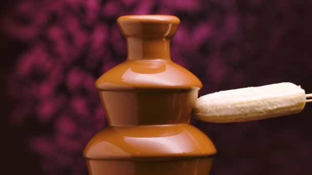 El chocolate con leche derretida fluye en una fuente de chocolate. El plátano maduro se sumerge y se envuelve en chocolate líquido caliente que desciende de las cascadas. Fondue. Fiesta y celebración. De cerca. Movimiento lento. — Vídeo de stock