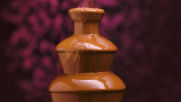 Chocolate fountain isolated on blurry purple background. Cocoa is poured onto the melted liquid dessert and dissolves in the liquid. Sweet yummy. Festive decoration. Close up. Slow motion. — Stock Video