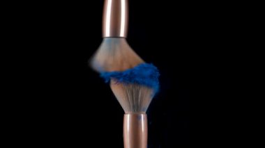 İki makyaj fırçası çarpışıyor ve siyah zemin üzerinde mavi toz parçacıklarının girdabına neden oluyor. Renkli gölgelerin izole edilmiş küçük parçacıkları uzayda uçar. Kapatın. Yavaş çekim.