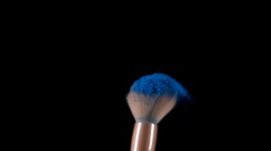 Yumuşak makyaj fırçası ürker ve siyah arkaplana karşı küçük mavi toz parçacıkları patlamasına neden olur. Renkli makyaj boyası uzaya doğru süzülür. Kapatın. Yavaş çekim.