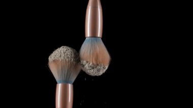 İki makyaj fırçası çarpışıyor ve siyah zemin üzerinde beyaz toz parçacıklarının girdabına neden oluyor. Ezilmiş kozmetik tozunun ince tozu güzel bir şekilde havada uçuşuyor. Kapatın. Yavaş çekim.