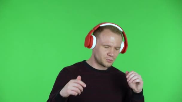 Retrato de un hombre de mediana edad bailando y disfrutando de la música rápida en grandes auriculares rojos. Hombre sin afeitar en un suéter rojo negro posando en una pantalla verde en el estudio. De cerca.. — Vídeos de Stock