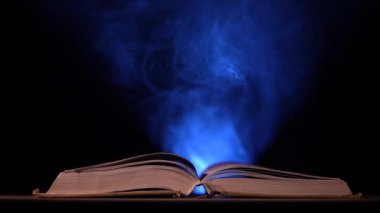 Karanlık, dumanlı bir stüdyoda, kalın, açık bir kitap siyah bir arkaplana karşı bir masanın üzerinde uzanıyor. Projektör ışığı altındaki toz parçacıkları açık bir kitabın üzerinde uçar. Ağır çekim. Kapat..
