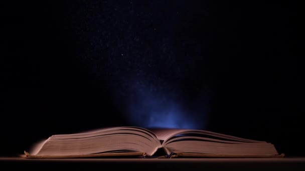 Ein aufgeschlagenes Buch fällt auf einen Tisch in einem dunklen Atelier. Ein Lichtstrahl von einem Suchscheinwerfer erhellt seine Seiten und die Staubpartikel, die darüber fliegen. Zeitlupe. Nahaufnahme. — Stockvideo