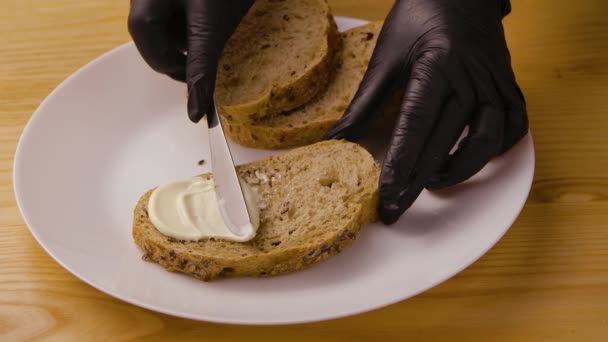 Руки в чорних рукавичках розмазують ножем на м'якому сирі або маслі на свіжому шматочку хліба. Прикрашає бутерброд на білій тарілці. Концепція ранкового сніданку перед робочим днем. Близько. Повільний рух . — стокове відео