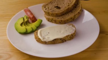 Siyah eldivenli eller tereyağlı taze ekmeğin üzerine jamon ve avokado dilimleri koyar. Beyaz tabakta iştah açıcı bir sandviç. İş gününden önce sabah kahvaltısı kavramı. Kapatın. Yavaş çekim.