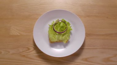 Farklı malzemelerden sandviç yapma ve yeme süreci. Hareket animasyonunu durdur. Üst Manzara. Taze ekmek, marul, soğan halkaları, domates, jamon ve peynir beyaz tabakta görünür. Kapat..