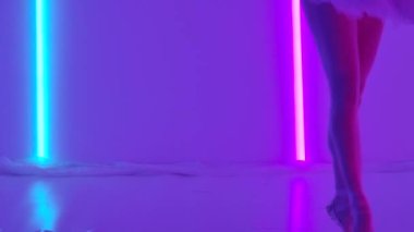 Zarif bir balerin, beyaz etekli klasik bir bale ve parlak neon tüplerin arka planında sivri uçlu ayakkabılar ile dans ediyor. Parmak ucunda dans eden bir dansçının ince bacakları. Yavaş çekim.
