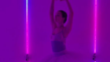 Kadın bale yapıyor ve stüdyoda neon ışıklarının arka planına karşı koreografik elementler sergiliyor. Kız bacağını dikey bir şekilde kaldırıyor ve dönüşleri ayarlıyor. Ağır çekim. Kapat..