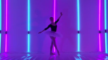 Profesyonel bir balerin, stüdyoda parmak uçlarında, parlak mavi ve pembe neon tüplerin arka planına karşı dans adımları atar. Klasik bir ablet koreografisi. Yavaş çekim.