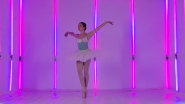Sahne elbisesi giymiş bir balerin atlayışlar, dikey bölünmeler ve diğer bale hareketleri yapar. Stüdyoda parlak neon lambaların arka planında profesyonel dansçı dans ediyor. Yavaş çekim.
