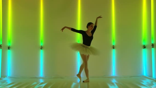 Uma jovem bailarina graciosa feminina está dançando elementos do balé clássico no estúdio contra um fundo de tubos de néon brilhantes. Menina em um tutu de balé e sapatos pontiagudos. Fechar. — Vídeo de Stock
