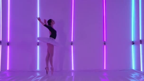 Young flexible ballerina in a white ballet tutu and pointe shoes in the studio against the background of neon tubes. The dancer performs a graceful dance of elements of classical ballet. Close up. — Stock Video