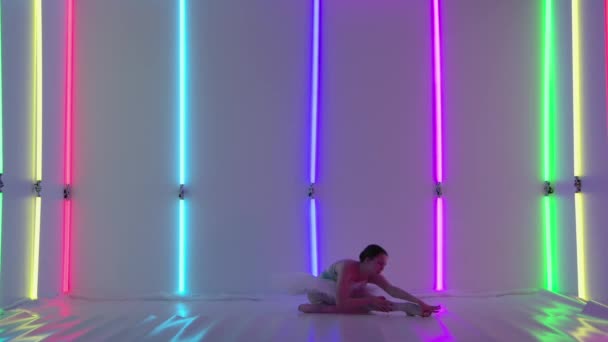 Ballerina in een witte tutu demonstreert haar flexibiliteit in de studio tegen een achtergrond van kleurrijke neon lichten. De danser, zittend op de vloer, maakt een split en staat dan op de brug. Sluiten.. — Stockvideo