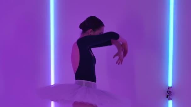 Een jonge slanke ballerina in een witte tutu voert sierlijke pas uit met armen en benen. Studio repetitie tegen de achtergrond van multi-gekleurde neon lichten. Langzame beweging. Sluiten.. — Stockvideo
