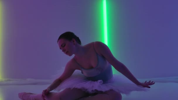 Ballerina démontre sa flexibilité en studio sur fond de néons colorés. La danseuse, assise sur le sol, travaille pas avec ses mains et fait une scission. Au ralenti. Gros plan. — Video