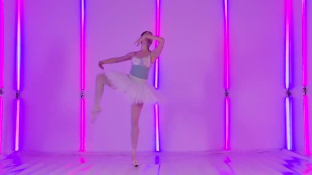 Una elegante bailarina está bailando un ballet clásico en el estudio sobre el fondo de brillantes lámparas de neón. La chica realiza divisiones verticales, pasos de baile y rotaciones. Movimiento lento. — Vídeos de Stock