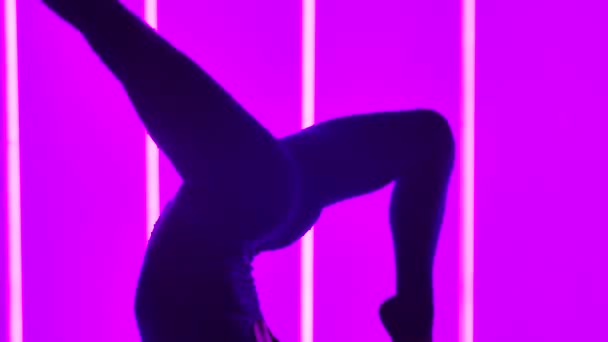 Giovane donna che fa esercizi ginnici in studio sullo sfondo di tubi al neon multicolore. Primo piano di una ragazza gambe in movimento in aria, che esegue una postazione. Silhouette. Rallentatore. — Video Stock
