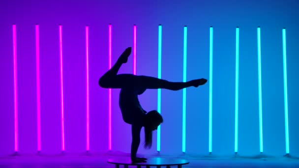 若い女性の体操選手がハンドスタンドをやっています。ピンクと青のネオン管を背景にスタジオで体操選手のシルエット. — ストック動画