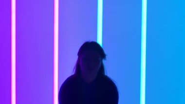 Stüdyodaki jimnastikçi, arka plandaki pembe ve mavi neon ışıklarına karşı. Kız ellerini kaldırır ve bacaklarını kaldırır, hareket ettirir. Kamera baştan aşağı hareket ediyor. Ağır çekim. Kapat..