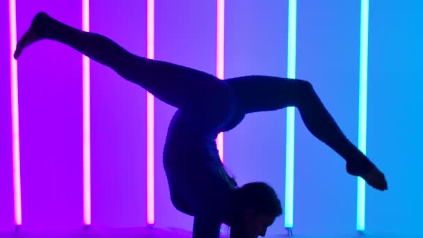 Vista lateral de una gimnasta haciendo un soporte de manos y moviendo elegantemente sus piernas en el aire. La chica en el estudio sobre un fondo rosa y azul. Primer plano de piernas de deportista. Silueta. Movimiento lento. — Vídeos de Stock