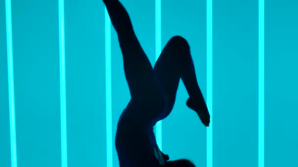 Pohled z boku na gymnastku, jak dělá stojku a pohybuje nohama ve vzduchu. Dívka je zapojena do studia na pozadí pestrobarevných neonových trubic. Silueta. Zpomal. Zavřít. — Stock video