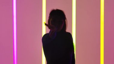 Stüdyodaki jimnastikçi çok renkli neon lambaların arka planına karşı. Oturma pozisyonunda bir kız, kollarının üzerinde durur ve bacaklarını kaldırır ve sicim yapar. Siluet. Kapatın. Yavaş çekim.