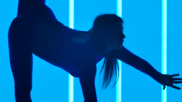 A gymnast raises her leg, leaning on her hands, bends her back and with one hand reaches for the raised leg in the studio against the background of neon tubes. Silhouette. Slow motion. Close up. — Stock Video