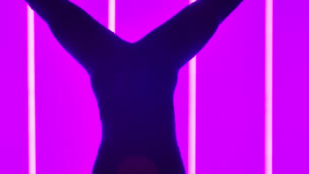 Gymnastka si užívá cvičení ve studiu na pozadí jasných neonových trubic. Dívka se během stojanu rozkročí a provádí pohyby nohama. Silueta. Zpomal. Zavřít. — Stock video