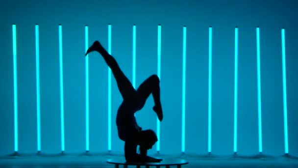 De atleet voert gymnastiek tegen de achtergrond van neon verlichting. Het meisje voert een handstand, op de borst, demonstreert de bewegingen van haar benen in de lucht. Donker silhouet. Langzame beweging. — Stockvideo